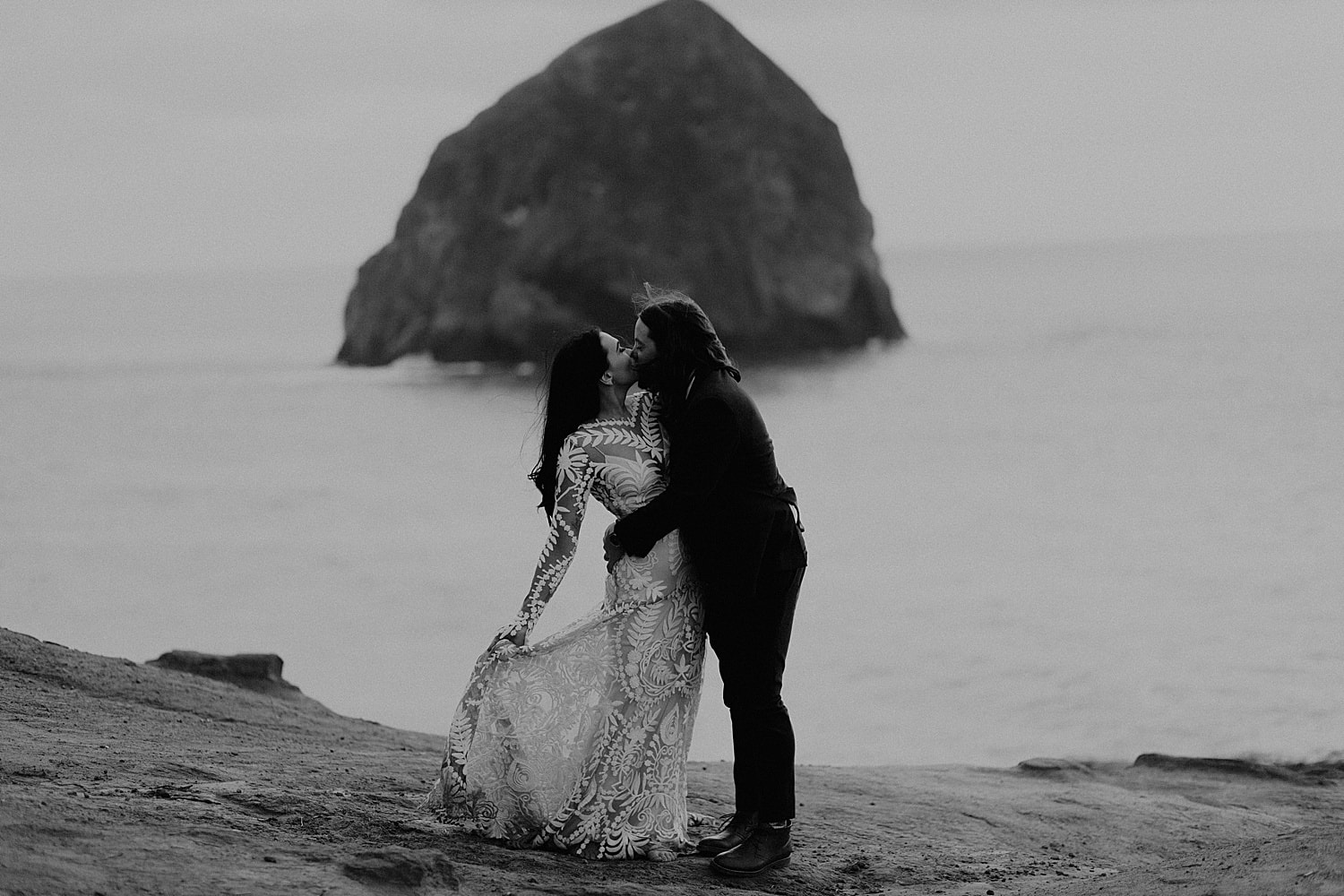black and white photo of a bride and groom kissing on the cliffside overlooking the ocean at Cape Kiwanda as they elope in Oregon