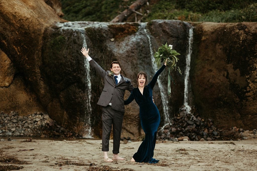 newlywed couple celebrating at a waterfall blue velvet wedding dress oregon coast hug point elopement captured by marcela pulido photography portland oregon wedding and elopement photographer