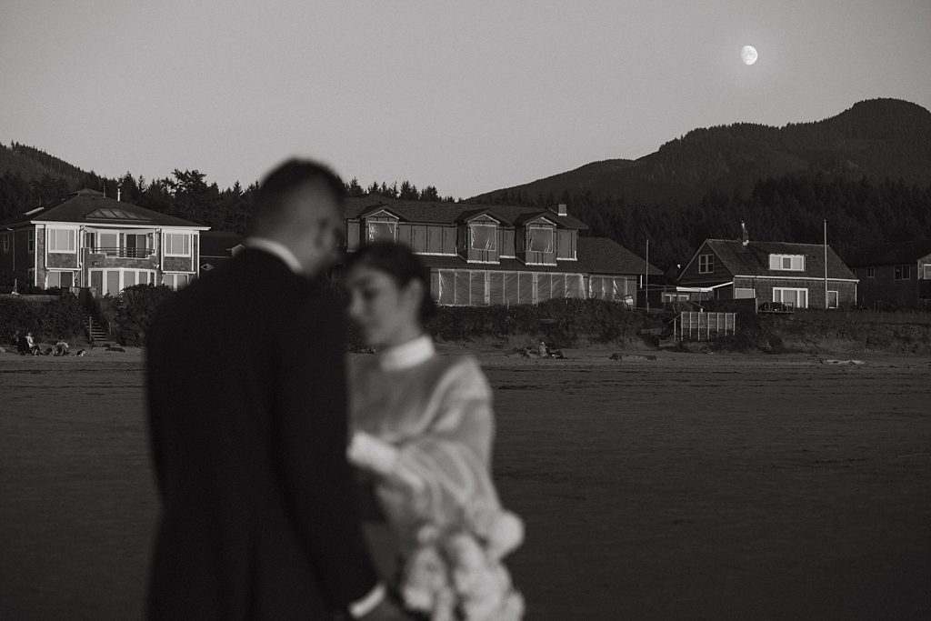 black and white cinematic portrait of newlywed couple with moon in the background at cannon beach elopement captured by marcela pulido portland oregon wedding photographer
